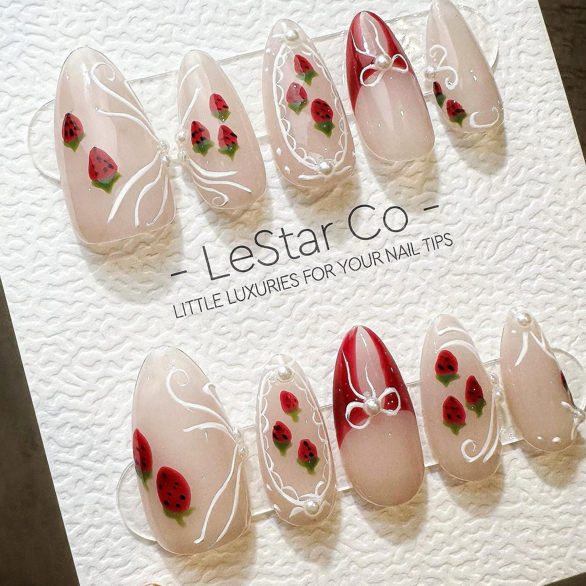 A set of ten almond-shaped press-on nails featuring a sheer nude base with hand-painted red strawberries, white swirling line accents, and small white pearl embellishments. The display card reads LeSt