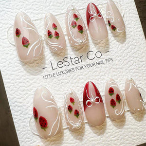 A set of ten almond-shaped press-on nails featuring a sheer nude base with hand-painted red strawberries, white swirling line accents, and small white pearl embellishments. The display card reads LeSt