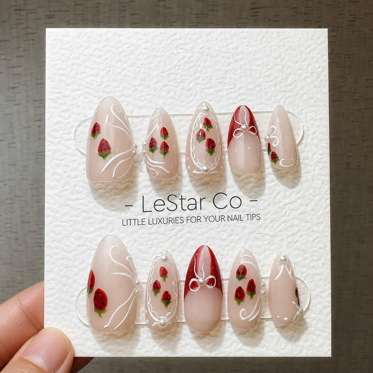 A set of ten almond-shaped press-on nails on a white display card labeled LeStar Co. The nails feature a nude base with hand-painted red strawberries, white swirling line accents, red tips, and small 