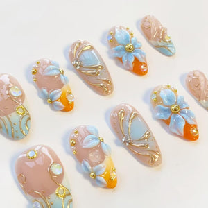 A set of ten press-on nails featuring a nude base with orange and light blue gradients. The designs include three-dimensional light blue flower petals, gold metallic swirls, small gold beads, and iridescent rhinestones on each nail.