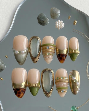 A set of ten almond-shaped press-on nails featuring a nude base with decorative accents including gold metallic bands, small white pearls, green resin elements, and brown tortoiseshell patterns, arranged on a reflective surface.
