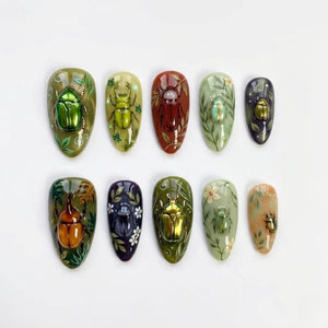 A set of ten almond-shaped press-on nails featuring three-dimensional metallic beetle embellishments. Each nail displays unique hand-painted botanical motifs, including leaves and flowers, in shades of green, brown, and purple.