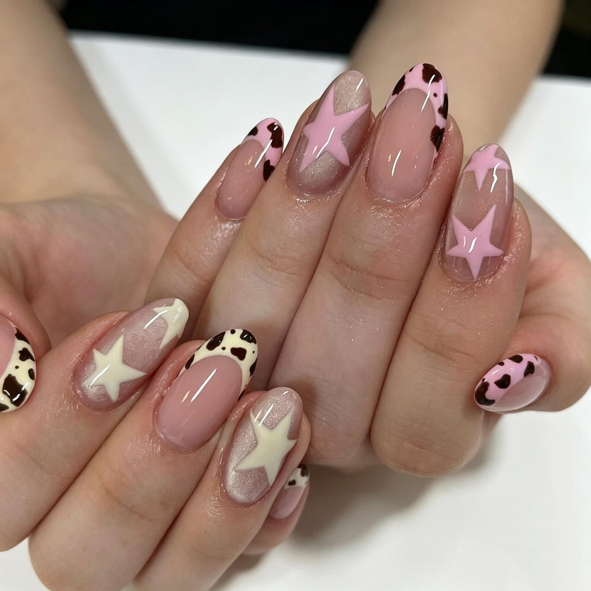 Oval-shaped nails featuring a mix of designs including light pink and cream-colored star motifs, French tips with brown and white cow print patterns, and a glossy finish over a natural-toned base coat on all ten fingernails.