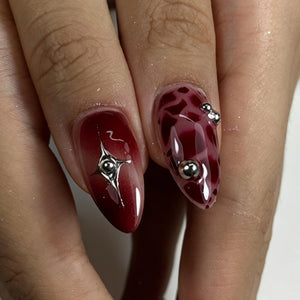 Two almond-shaped fingernails featuring a deep red gradient polish. One nail displays a central silver star-shaped charm with a small sphere, while the other features a dark red marbled pattern accented with three small silver metallic spheres.