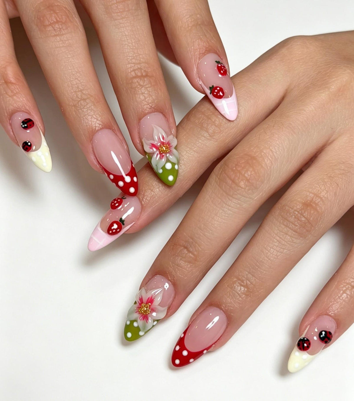 Almond-shaped nails featuring a French manicure style with a nude base. The tips are decorated with red and white polka dots, green and white polka dots, and cream colors. Accents include small red strawberries and ladybugs.