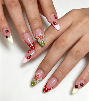 Almond-shaped nails featuring a French manicure style with a nude base. The tips are decorated with red and white polka dots, green and white polka dots, and cream colors. Accents include small red strawberries and ladybugs.