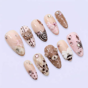 A set of ten almond-shaped press-on nails featuring a light pink and brown color palette. The designs include 3D bear faces, white bows, floral patterns, musical notes, stars, polka dots, and small clear rhinestones on a glossy finish.