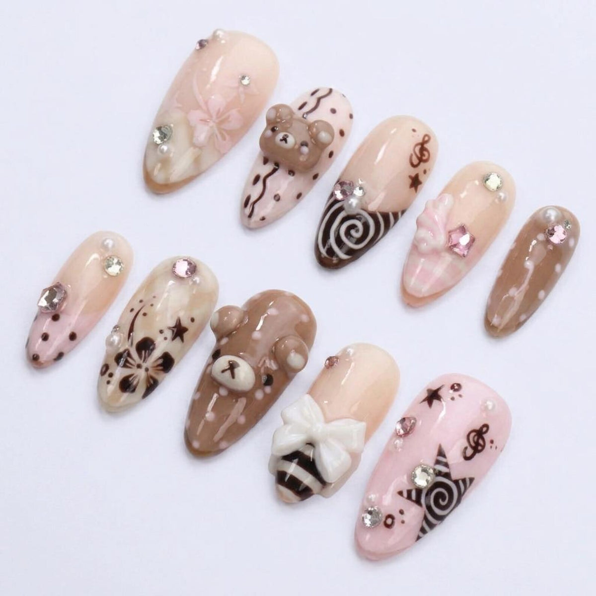 A set of ten almond-shaped press-on nails in shades of beige, brown, and soft pink. The designs feature 3D bear figures, white bows, brown floral patterns, musical notes, stars, polka dots, and small clear rhinestones on each nail.