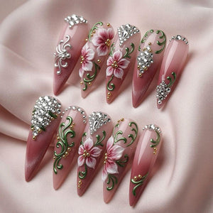 A set of ten stiletto-shaped press-on nails featuring a pink ombre base. The designs include 3D white and pink floral accents, metallic green swirls, small gold beads, and clusters of clear rhinestones arranged in various patterns.