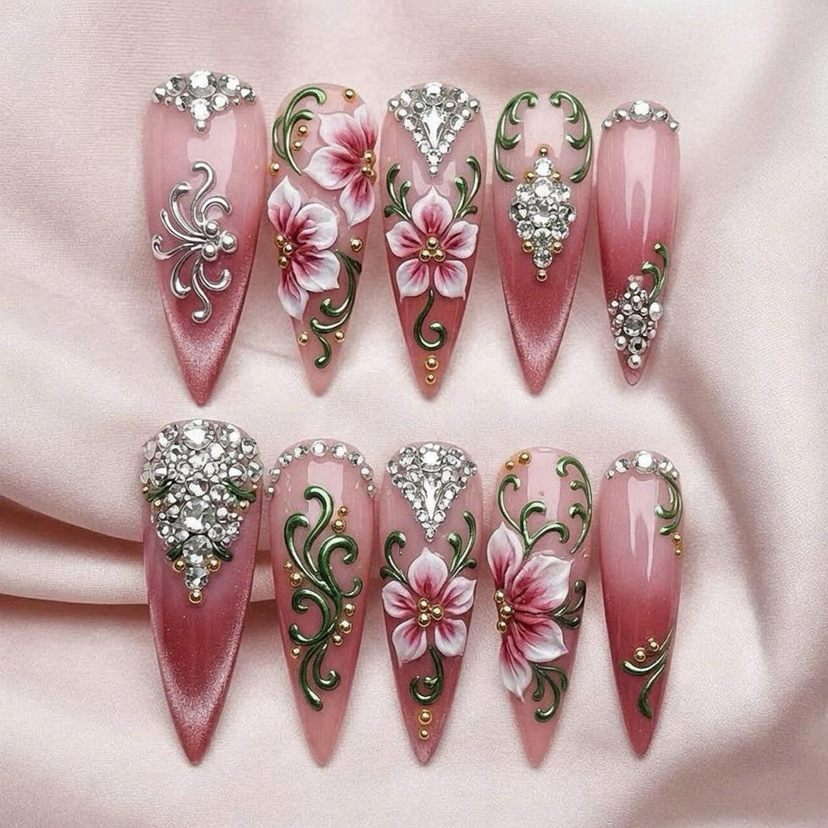 Ten stiletto-shaped press-on nails featuring a pink gradient base. The designs include hand-painted white and pink flowers, green swirling vines, gold metallic accents, and clusters of clear rhinestones arranged in various patterns.
