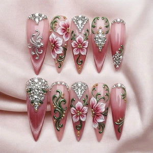 Ten stiletto-shaped press-on nails featuring a pink gradient base. The designs include hand-painted white and pink flowers, green swirling vines, gold metallic accents, and clusters of clear rhinestones arranged in various patterns.
