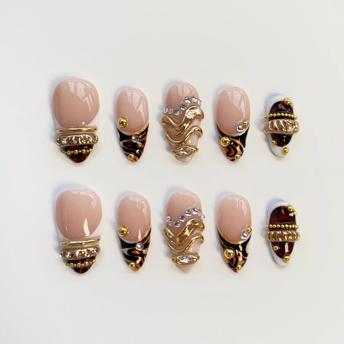 A set of ten press-on nails featuring a nude base with tortoise shell patterns, gold metallic accents, and clear rhinestones. The designs include horizontal bands, wavy lines, and small gold spheres arranged on almond-shaped tips.