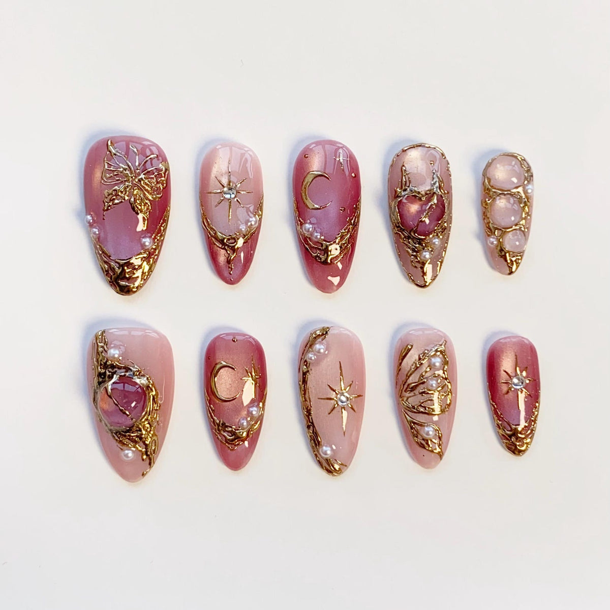 A set of ten press-on nails in varying shades of pink, featuring gold metallic relief designs, small white pearls, and embedded crystals. The patterns include butterflies, crescent moons, and starbursts on almond-shaped tips.