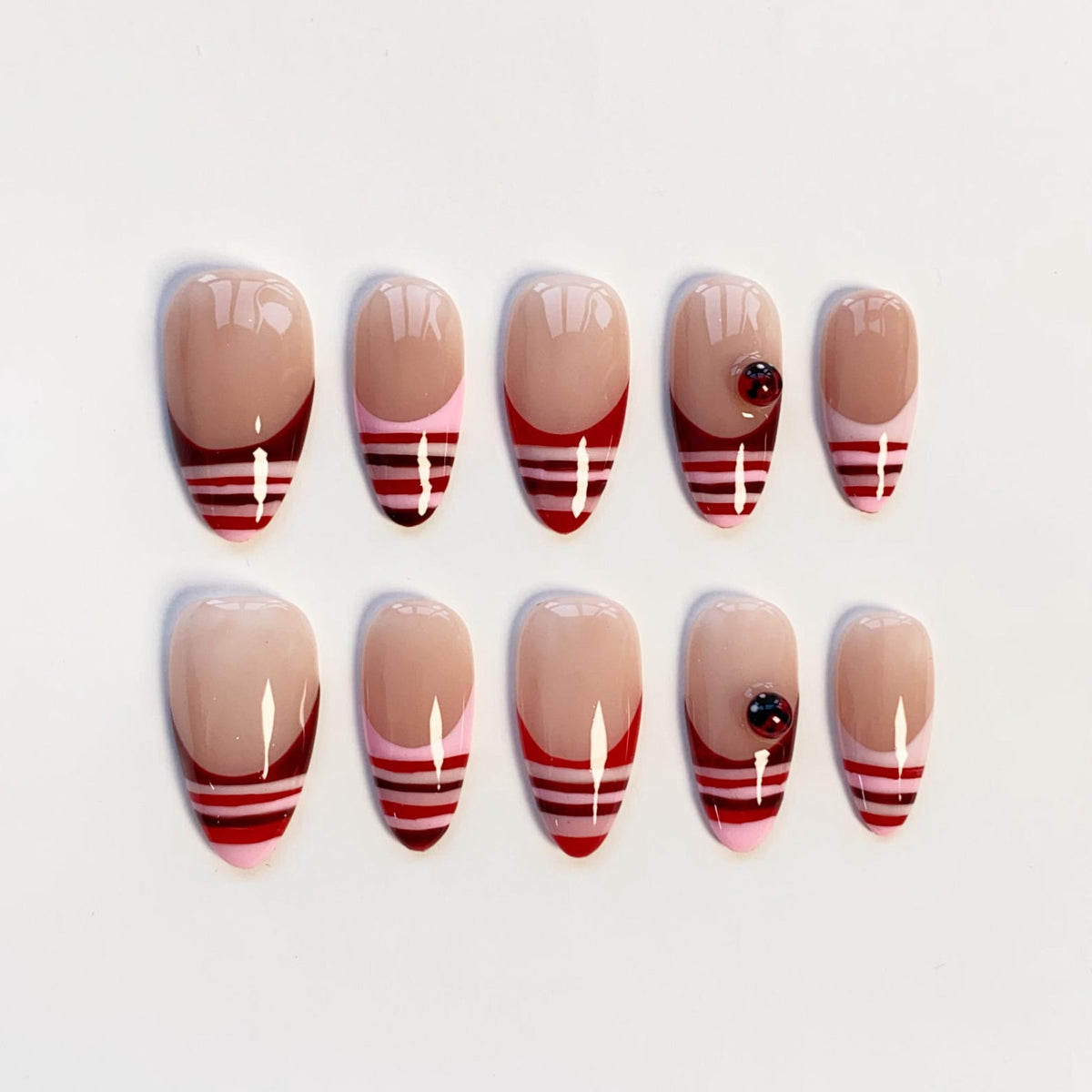 A set of ten almond-shaped press-on nails featuring a nude base with French tips. The tips are decorated with horizontal stripes in shades of red, dark red, and light pink. Two of the nails feature a small, round, dark red gem.