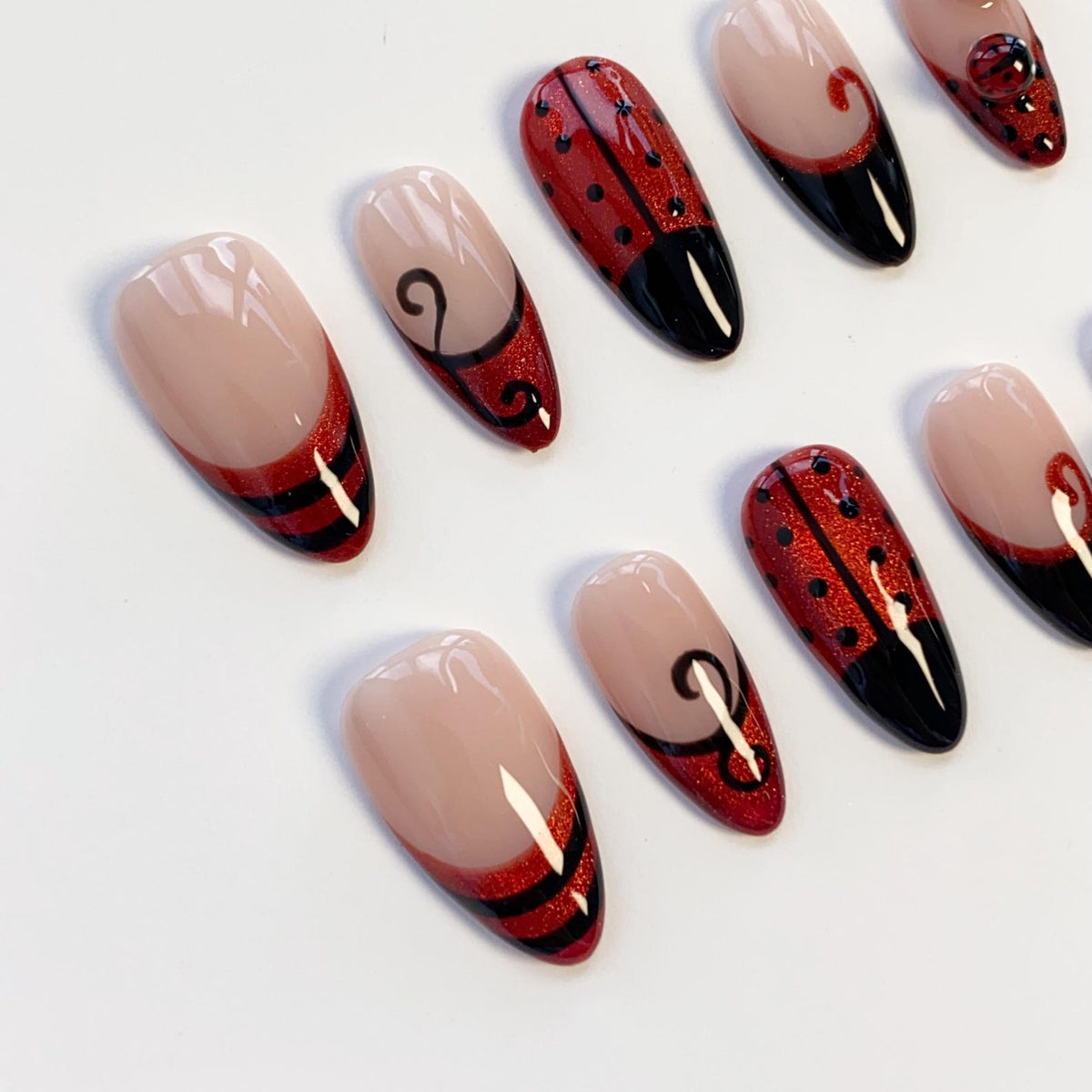 A set of almond-shaped press-on nails featuring a nude base with various designs including red glitter ladybug patterns with black spots, black French tips, and black swirl accents. The nails have a glossy finish and are arranged in a group.