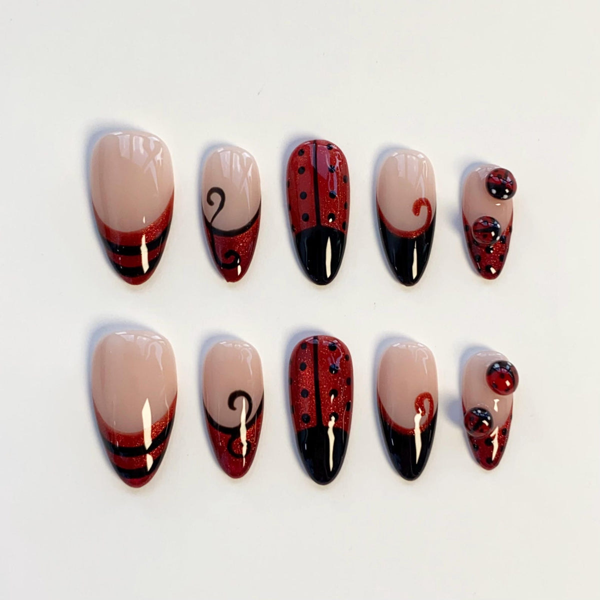 A set of ten almond-shaped press-on nails featuring a nude base with red and black designs. The patterns include horizontal stripes, black swirls, ladybug-inspired red glitter with black dots, and small raised red and black spheres.