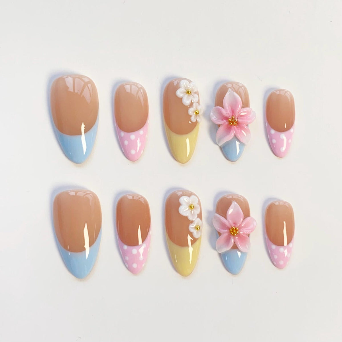 A set of ten almond-shaped press-on nails featuring a nude base with pastel tips in light blue, yellow, and pink with white polka dots. Two nails are adorned with three-dimensional white flowers, and two feature large pink flowers.