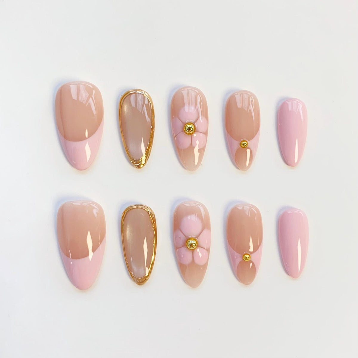 A set of ten almond-shaped press-on nails featuring a variety of designs including light pink French tips, clear nails with gold metallic edges, light pink floral accents with gold centers, and solid light pink nails with gold studs.