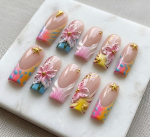 A set of ten press-on nails featuring a nude base with French tips. The designs include colorful abstract patterns in pink, yellow, and blue, three-dimensional pink floral accents, gold starfish charms, and small gold metallic studs.