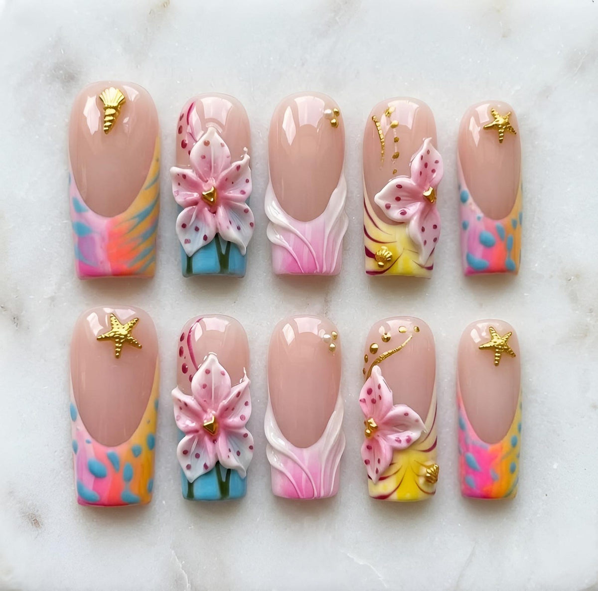 A set of ten press-on nails featuring a nude base with colorful French tips in shades of pink, blue, and yellow. The design includes three-dimensional pink floral accents, gold starfish and seashell charms, and small gold bead embellishments.