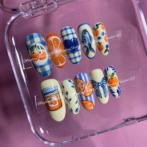 A set of ten press-on nails featuring various hand-painted designs including blue gingham patterns, orange slices, a blue heart, a fish, cherries, a jam jar, a blue bow, and botanical motifs. The text MaydayStudioUS is visible.