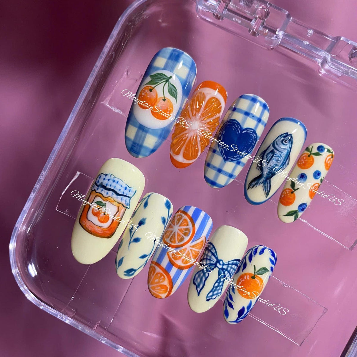 A set of ten press-on nails featuring various hand-painted designs including blue gingham patterns, orange slices, a blue heart, a fish, a jar of orange marmalade, blue bows, and small orange and blueberry motifs on cream backgrounds.