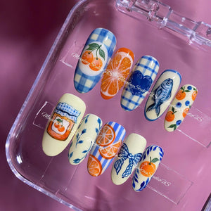A set of ten press-on nails featuring various hand-painted designs including blue gingham patterns, orange slices, a blue heart, a fish, a jar of orange marmalade, blue bows, and small orange and blueberry motifs on cream backgrounds.
