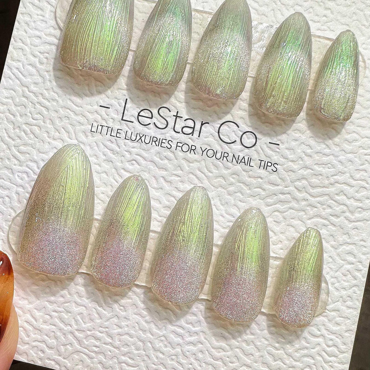 A set of ten almond-shaped press-on nails featuring a textured, iridescent green finish with a fine glitter gradient at the base. The nails are displayed on a white card labeled LeStar Co - LITTLE LUX