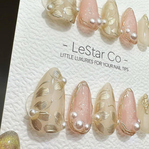 A set of press-on nails featuring a soft pink and translucent beige palette with gold leaf floral accents and small white pearls. The nails are displayed on a white card with the text LeStar Co - LITT
