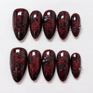 A set of ten almond-shaped press-on nails featuring a deep burgundy and black marbled base with intricate hand-painted dark red rose motifs. Some nails are accented with small, metallic silver spherical beads and thin lines.