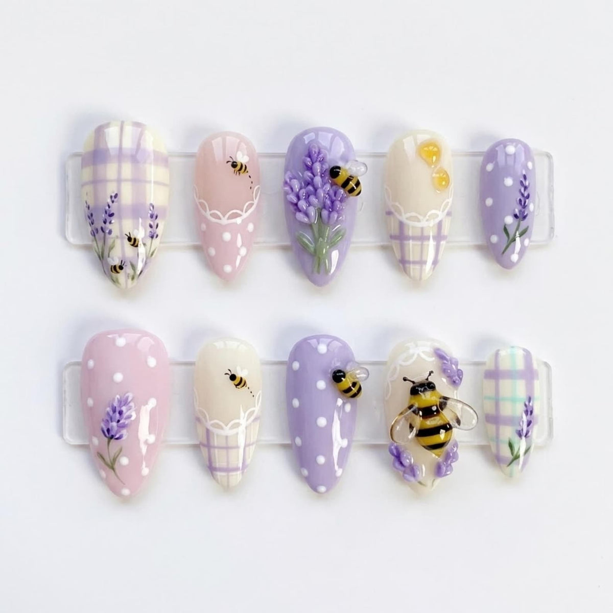 A set of ten almond-shaped press-on nails featuring lavender, white, and cream colors. Designs include lavender sprigs, small bees, white polka dots, and plaid patterns. One nail features a large, three-dimensional bee embellishment.