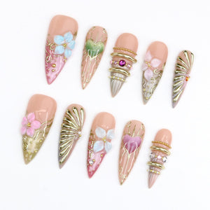 A set of ten stiletto-shaped press-on nails featuring a nude base with intricate 3D designs. Embellishments include gold metallic accents, raised pink and blue floral motifs, heart-shaped patterns, and small embedded rhinestones.