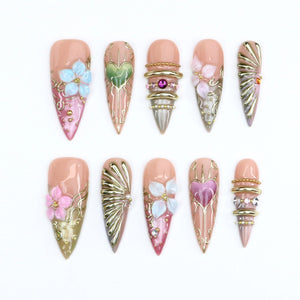 A set of ten stiletto-shaped press-on nails featuring a nude base with intricate 3D embellishments. Designs include raised light blue and pink flowers, metallic gold filigree, heart motifs, stacked gold rings, and small rhinestones.