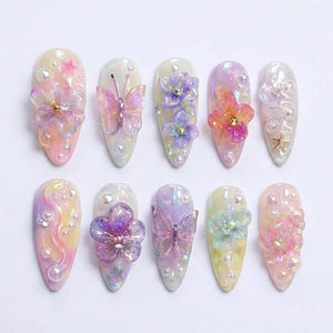 A set of ten almond-shaped press-on nails featuring a pastel iridescent finish. The designs include three-dimensional acrylic butterflies, purple and pink floral accents, small white pearls, and tiny gold star charms arranged on each nail.