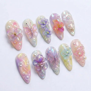 A set of ten almond-shaped press-on nails featuring a pastel iridescent finish. The designs include three-dimensional translucent flowers, butterflies, small white pearls, and tiny gold star accents arranged across the nail surfaces.