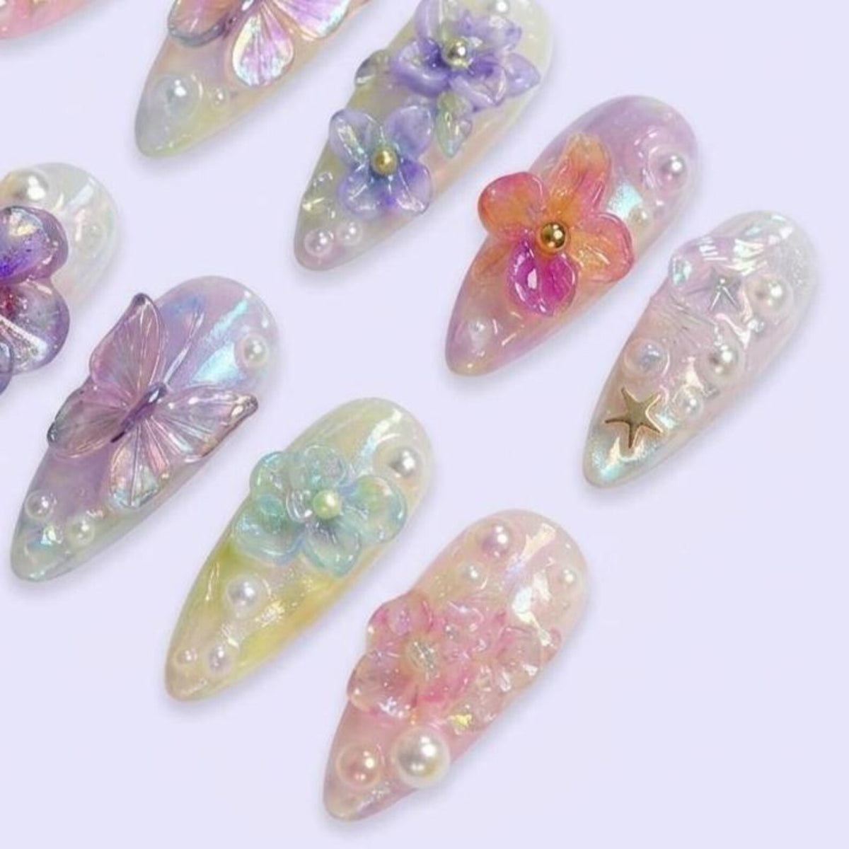 A collection of almond-shaped press-on nails featuring iridescent finishes, 3D floral and butterfly embellishments, and small white pearls. Some nails include gold-toned star accents, while others display translucent petals in shades of pink, purple,