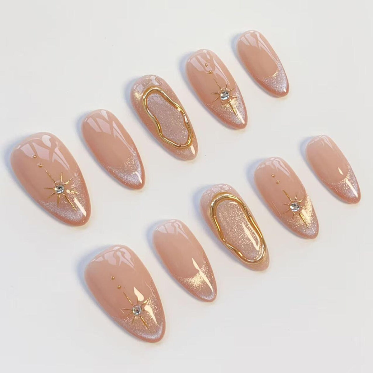 A set of ten almond-shaped press-on nails in a nude base color. The design features a shimmering finish, gold metallic accents in wavy line patterns, and starburst motifs centered with small clear rhinestones on select nail tips.