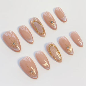 A set of ten almond-shaped press-on nails in a nude base color. The design features a shimmering finish, gold metallic accents in wavy line patterns, and starburst motifs centered with small clear rhinestones on select nail tips.