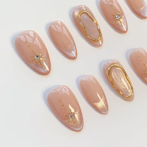 A set of almond-shaped press-on nails in a nude base color. The design features gold starburst accents with small clear rhinestones, gold metallic wavy line details, and subtle shimmering glitter gradients on the tips of the nails.