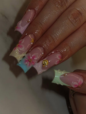Long square-shaped artificial nails featuring a French manicure style with pastel-colored tips in blue, pink, and green. The nails are decorated with hand-painted pink floral designs, small white pearl accents, and a gold seashell charm.