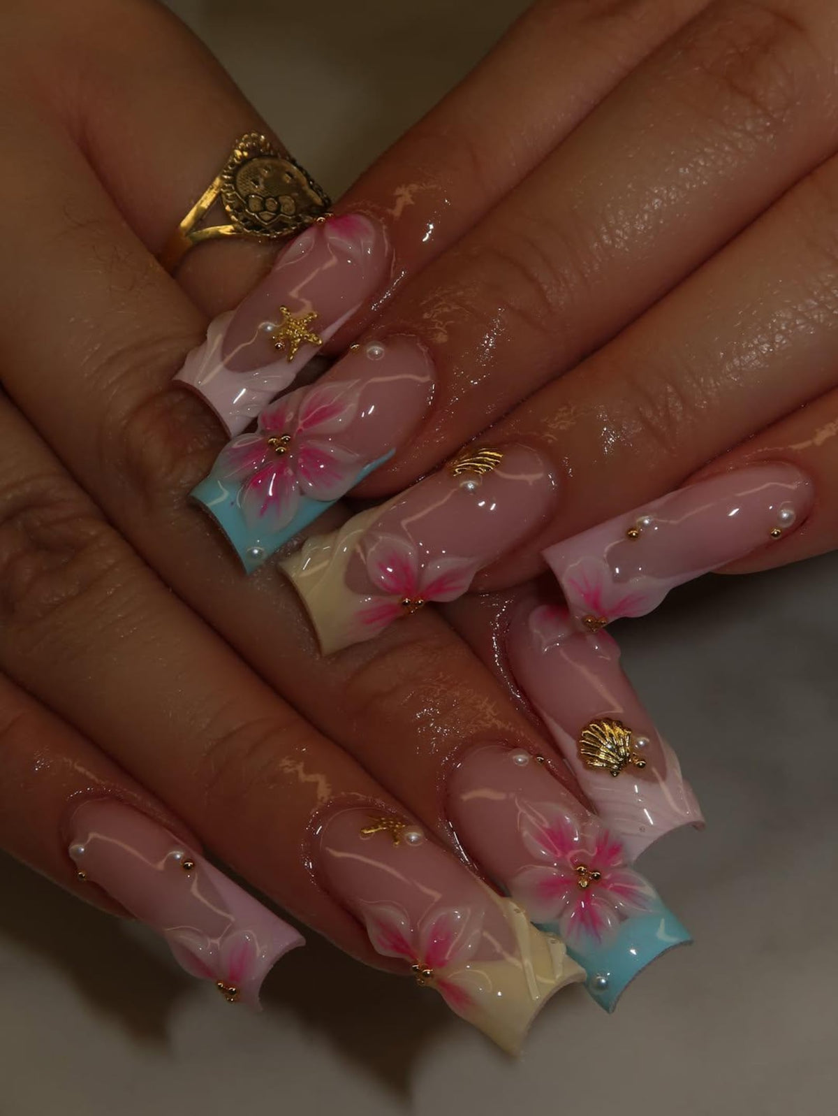 Long square-shaped artificial nails featuring a French manicure style with pastel blue, yellow, and pink tips. The nails are decorated with hand-painted pink floral designs, small gold-toned starfish and seashell charms, and tiny white pearls.