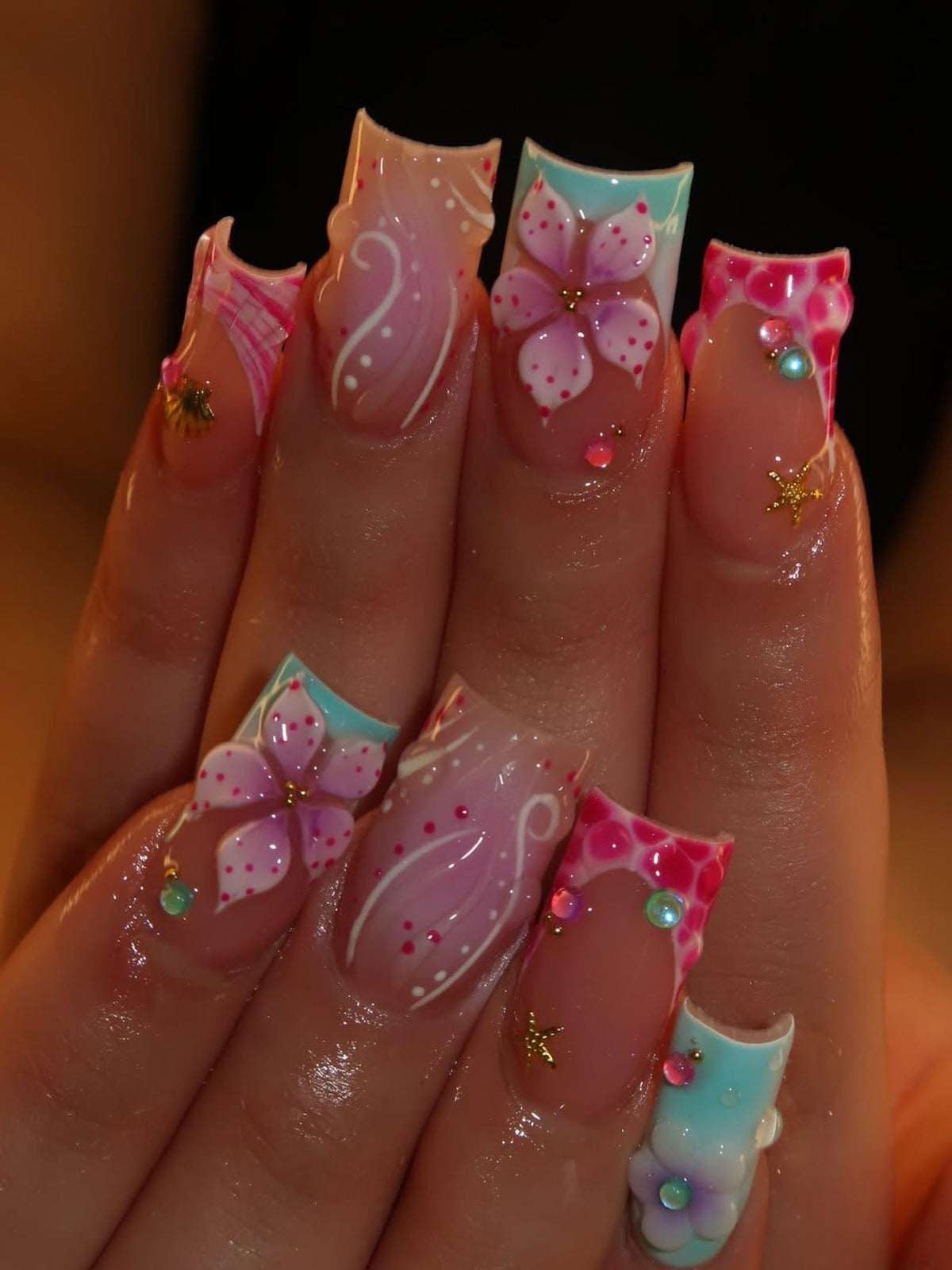 Square-shaped artificial nails featuring a mix of designs including pink and white floral patterns, swirling white lines, and bright pink spotted textures. The nails are decorated with small gold starfish charms and colorful rhinestones.