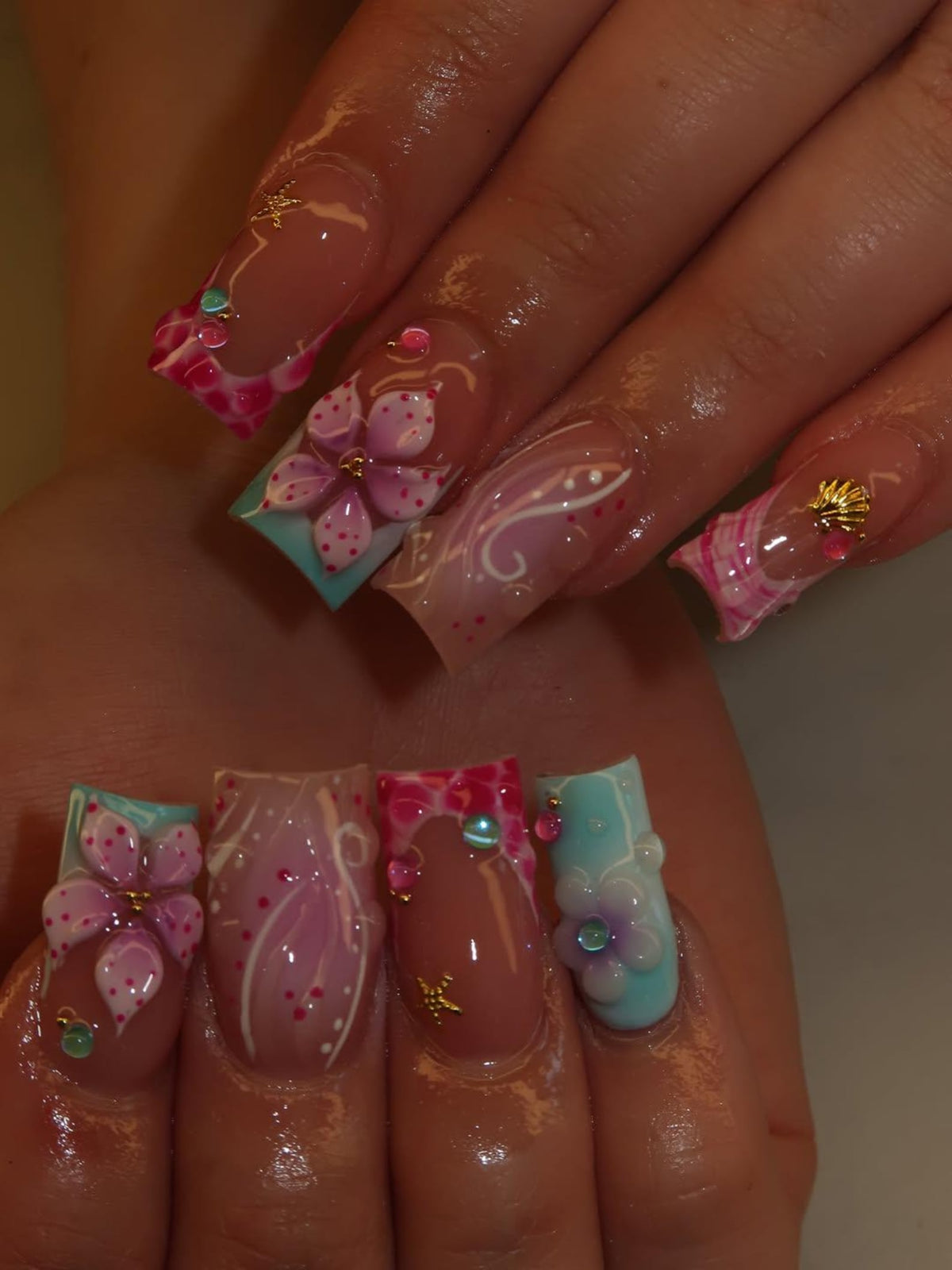 Square-shaped artificial nails featuring a mix of designs including pink and purple 3D floral accents, light blue and pink gradients, white swirl patterns, small gold starfish and seashell charms, and tiny colorful rhinestones.