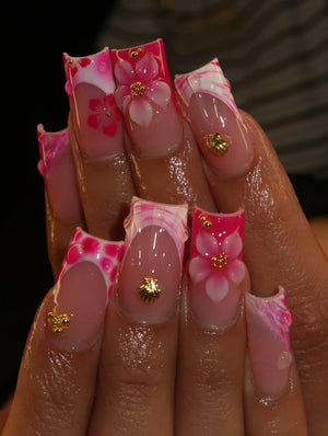 Long square-shaped artificial nails featuring a pink and white French tip design. The nails are decorated with three-dimensional pink and white floral accents, small gold metallic shell charms, and tiny gold beads on a clear base.
