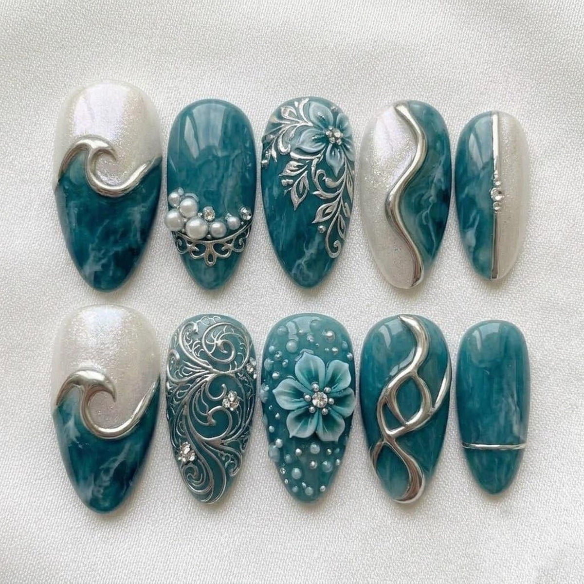 A set of ten press-on nails featuring a teal and white marble pattern. Designs include silver wave accents, white pearl embellishments, intricate silver floral filigree, a sculpted teal flower, and silver metallic lines on almond-shaped tips.