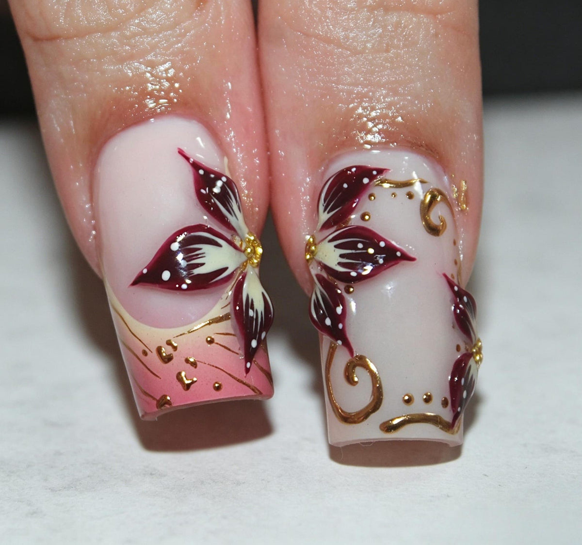 Two fingernails featuring a light pink base coat with hand-painted floral designs in deep burgundy and white. The artwork includes gold metallic swirls and small dots, with a gradient pink tip on one nail and intricate petals.