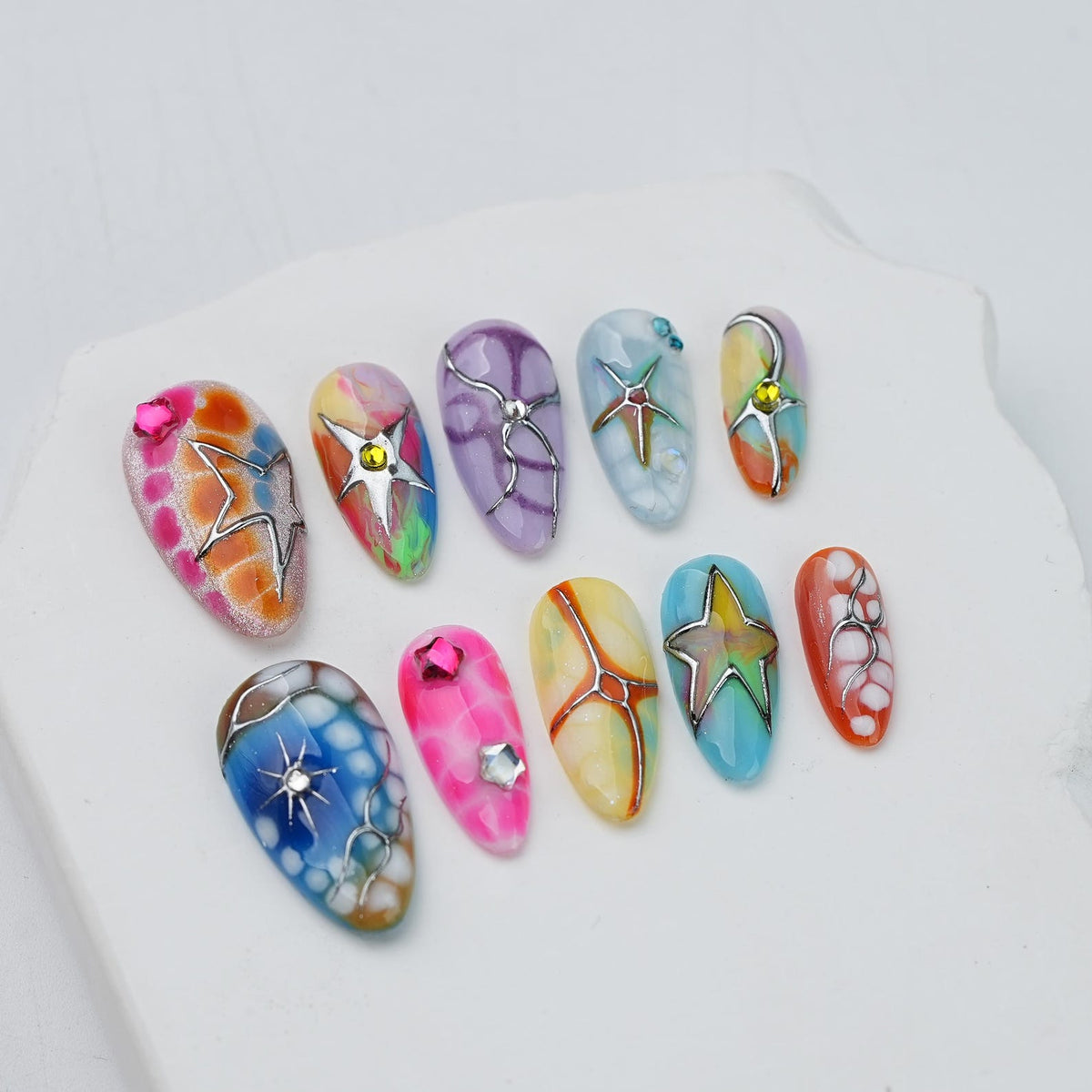 A set of ten almond-shaped press-on nails featuring colorful marbled patterns, metallic silver star and abstract line overlays, and small embedded rhinestones in various shapes and colors, arranged in two rows on a white surface.
