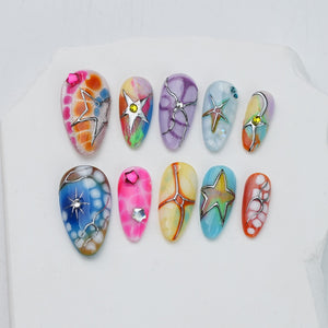 A set of ten press-on nails featuring diverse designs including colorful marbled patterns, metallic silver star outlines, and small embedded rhinestones in pink, yellow, and clear tones, arranged in two rows on a white surface.