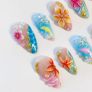 A set of press-on nails featuring tropical ocean themes, including hand-painted hibiscus flowers, starfish, dolphins, and seashells. The designs incorporate 3D elements like gold beads, pearls, and iridescent rhinestones on blue and nude bases.