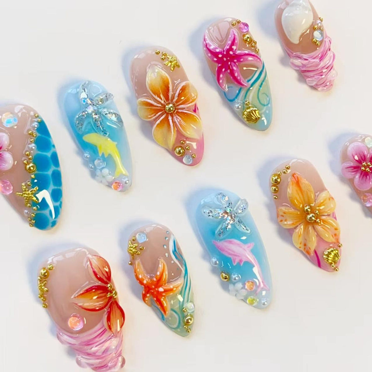 A set of ten almond-shaped press-on nails featuring tropical and ocean-themed designs. The nails are decorated with 3D floral motifs, starfish, dolphins, seashells, and small gold beads, painted in vibrant shades of pink, orange, and blue.